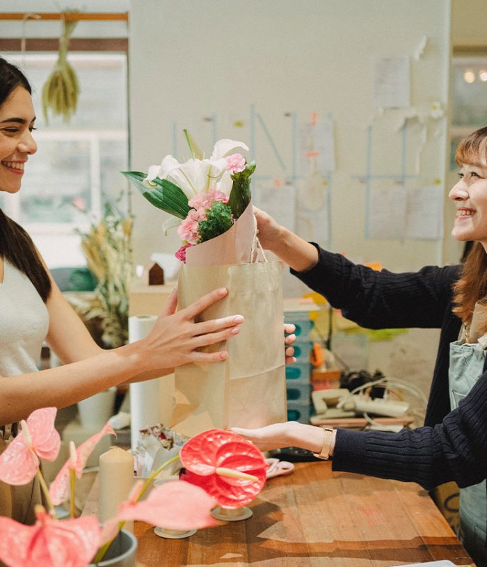 Asian retail worker handing over a white paper shopping bag filled with flowers to smiling customer on the other side of the counter